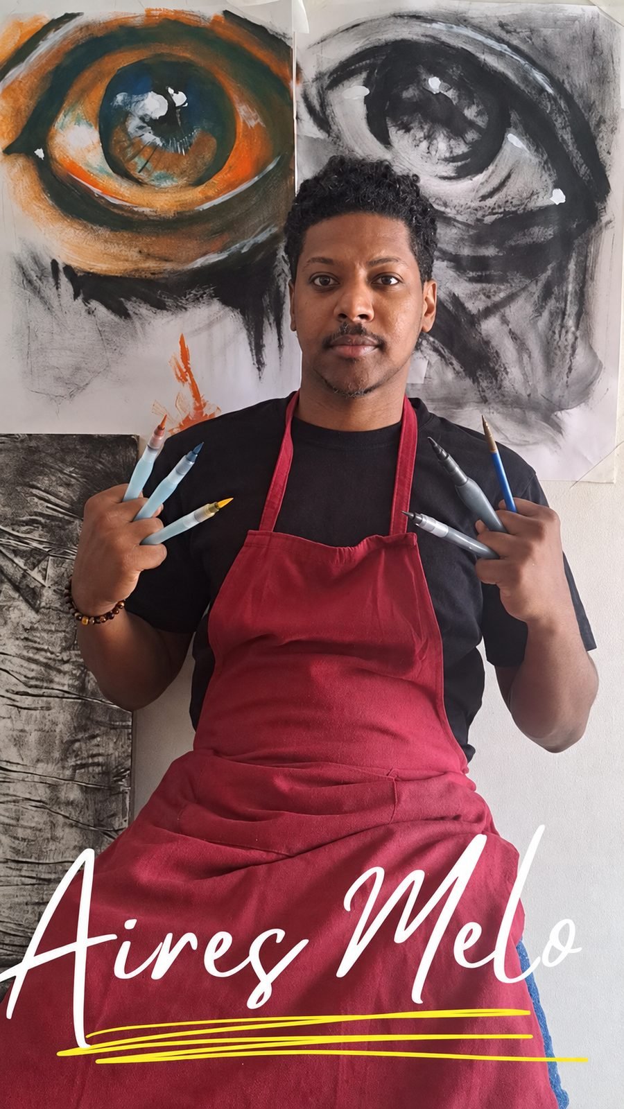 Aires Melo, Cape Verdean visual artist, holding his ink and charcoal tools in front of two of his eye studies.