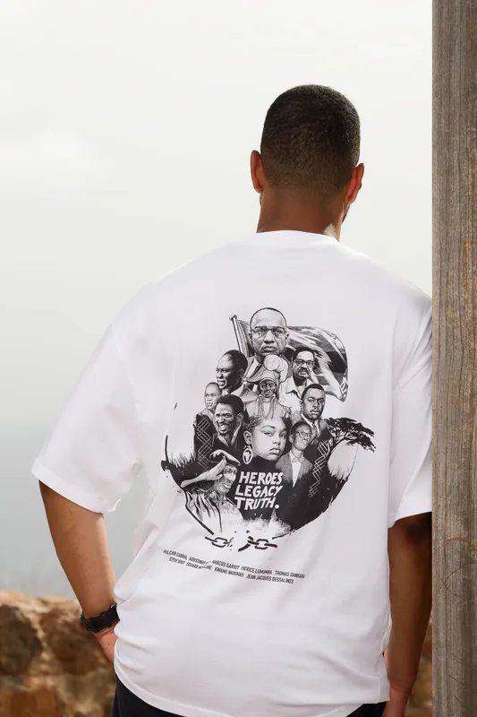 Back of the Fidju Ink shirt by a stone column overlooking the sea, Cabo Verde — the nine-heroes print.